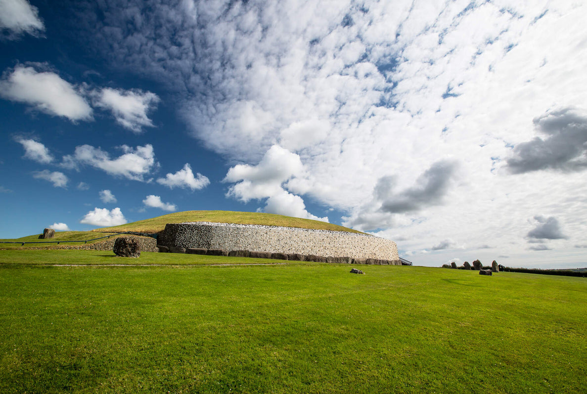 Newgrange monument sits on a green hillside under a partly cloudy sky. The landscape features rolling grass