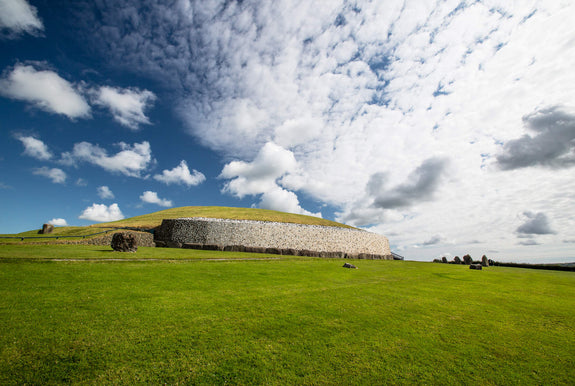 Newgrange monument sits on a green hillside under a partly cloudy sky. The landscape features rolling grass