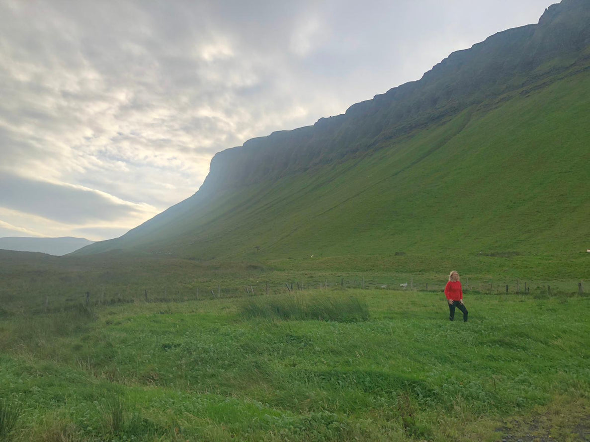 Lulu stands at the foot of Benbulben, County Sligo wearing a red Aran sweater under a cloudy sky. The landscape features rolling hills and a sense of openness.