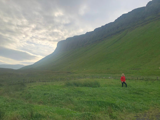 Lulu stands at the foot of Benbulben, County Sligo wearing a red Aran sweater under a cloudy sky. The landscape features rolling hills and a sense of openness.