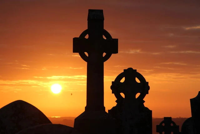 Silhouetted Celtic crosses stand against a vibrant sunset, with the sun dipping below the horizon. 