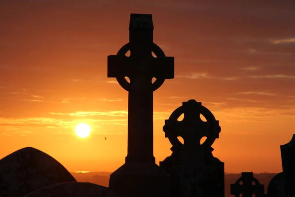 Silhouetted Celtic crosses stand against a vibrant sunset, with the sun dipping below the horizon. 