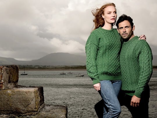 A man and woman stand side by side on a rocky shore, both wearing matching green Aran sweaters with a textured pattern. The background features a cloudy sky and distant mountains, with a body of water visible. 