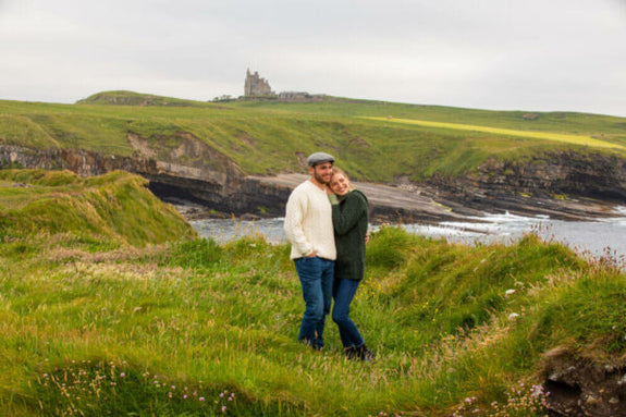 A couple stands together on a grassy hillside by the coast, smiling and embracing. Behind them, a cliffside overlooks the ocean, with a historic structure visible in the background. 