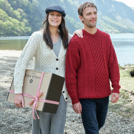 A woman in a cream-colored cable-knit cardigan and a man in a red cable-knit sweater walk by a lake. The woman is holding a gift box wrapped with pink ribbon, 