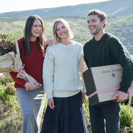 Three smiling individuals stand outdoors in a scenic landscape, each wearing cozy knitted Aran sweaters. The woman on the left carries a bouquet, while the man on the right holds a gift box with a ribbon. 