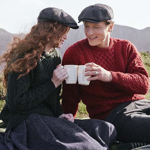 A couple dressed in cozy knit Aran sweaters and flat caps sit on a hillside, smiling and holding mugs. The landscape is mountainous, suggesting a serene, outdoor setting.