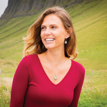 A woman with long hair and a bright smile wears a red sweater and silver Celtic jewelry with green stones, standing outdoors in front of a green hillside.