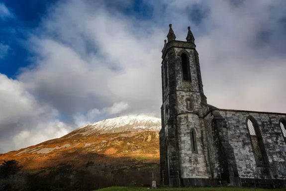 Poisoned Glen, Donegal. A stone church tower stands prominently against a backdrop of a snow-capped mountain under a partly cloudy sky.