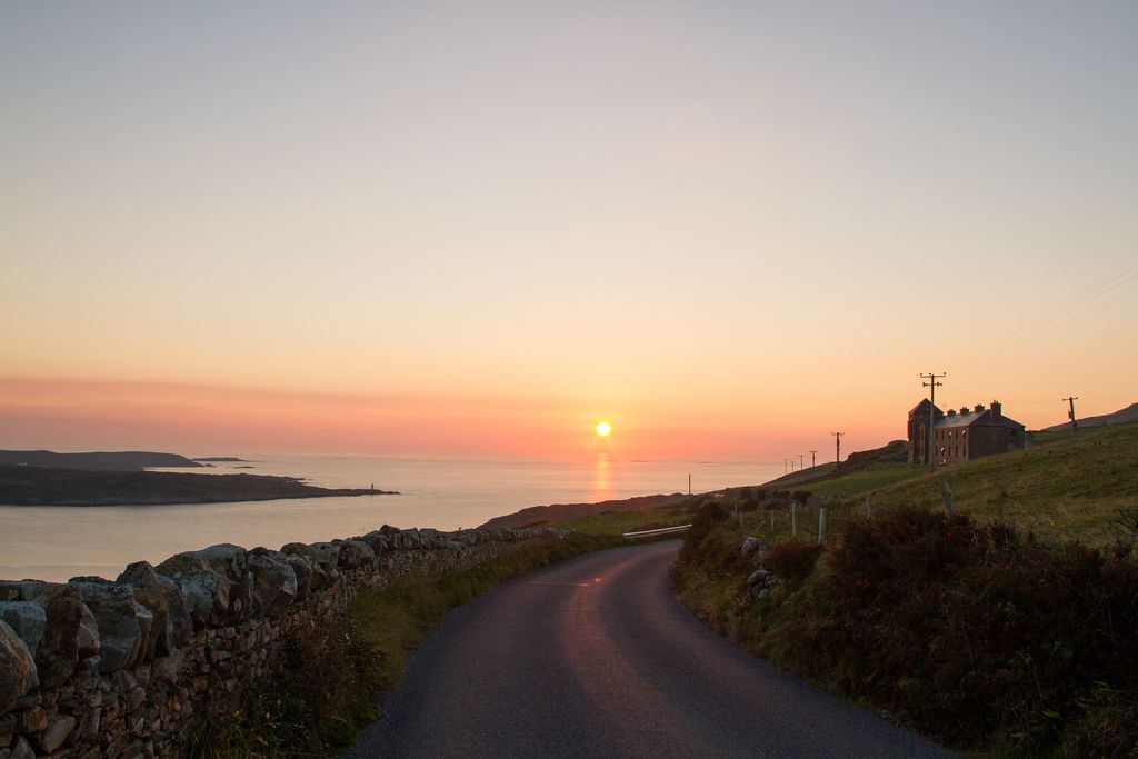 A winding Irish country road leads towards a coastal view at sunset, with the sun setting over the ocean in the background.