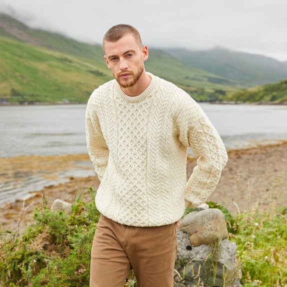 A person wearing a cream-colored cable knit sweater stands by a scenic lake and green mountains on a cloudy day.