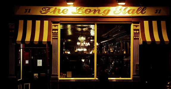 Exterior of The Long Hall pub in Dublin is illuminated at night. The window displays an interior with warm lighting creating an inviting atmosphere.