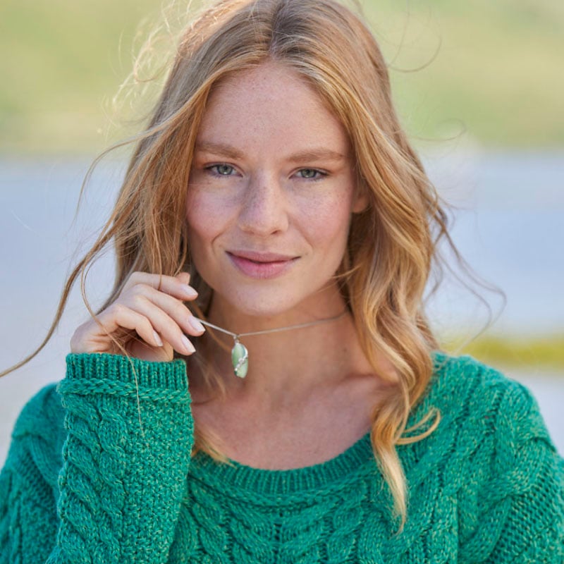 A person with long, wavy blonde hair wearing a green knitted sweater, holding a pendant necklace near their face. They are standing outdoors with a blurred natural background.
