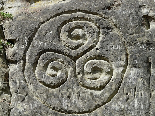 A carved stone featuring a triple spiral triskele motif, set against a rugged rock surface. The spirals are concentric, with each one closely intertwined.