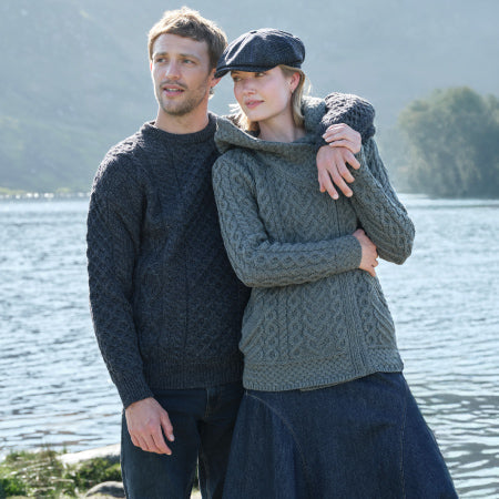 A man and a woman stand together by a lake, wearing cozy, knitted sweaters. The man has short hair and smiles at the camera while the woman leans slightly against him, sporting a cap. The background features trees and hills, suggesting an outdoor setting.