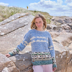 A woman standing by rock wearing a hand-knitted grey and white sheep patterned sweater.
