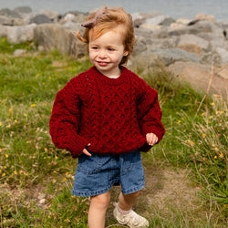 A young child walks on grass near a rocky shoreline, wearing a vibrant red kid's Aran sweater and blue denim shorts. The scene is set outdoors with greenery in the background and water visible in the distance.