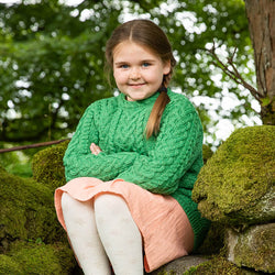 A young girl sits on moss-covered stones in a natural setting. She wears a green kids Aran sweater and a light pink skirt. Her arms are crossed, and she has a friendly smile. Trees and foliage create a lush background.