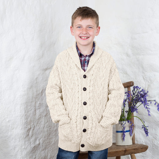 A young boy stands smiling, wearing a cream-colored kids Aran cardigan over a plaid shirt. He has his hands in the pockets of the cardigan and is positioned next to a wooden stool with a vase of purple flowers on it. The background features textured white walls.