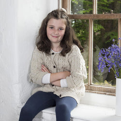 A young girl with curly hair sits on a windowsill, wearing a  beige kids Aran cardigan in 100% merino wool. She has her arms crossed and smiles softly at the camera. A vase of purple flowers is placed beside her, with greenery visible through the window behind her.