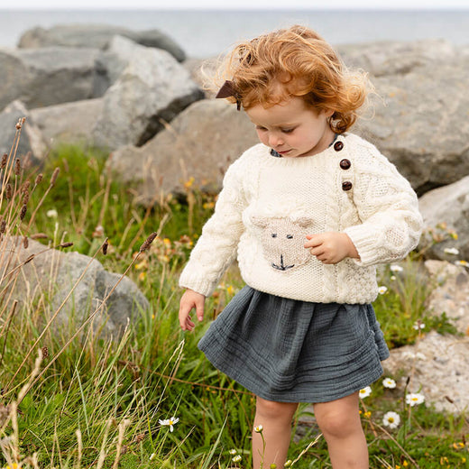 A young child stands among rocks and wildflowers by the sea. She wears a cozy kids Aran sweater with a sheep design and buttons, paired with a dark skirt. The scene captures a natural, playful moment on a grassy shoreline.