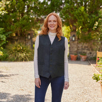 Woman wearing a dark gray tweed vest outdoors with greenery in the background