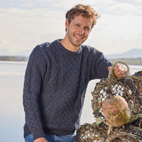 Man wearing a dark blue Aran  sweater holding fishing nets by a body of water.