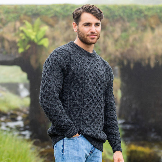 Man wearing a dark gray knitted sweater standing outdoors with greenery in the background