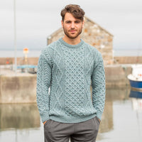 A man stands confidently in front of a harbor, wearing a light blue cable-knit Aran  sweater and grey trousers. The background features boats and a stone building under an overcast sky.