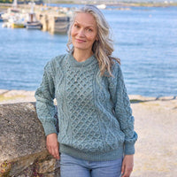Woman wearing a pale blue Aran sweater by a waterfront with boats in the background. 