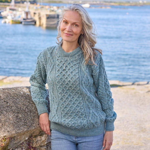 Woman wearing a pale blue Aran sweater by a waterfront with boats in the background. 