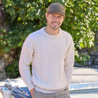 Man wearing a cream Aran sweater and cap standing outdoors with greenery in the background