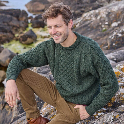 A man sits on rocky terrain by the water, wearing a dark green Aran sweater with a textured pattern and tan pants. He smiles as he gazes off to the side, surrounded by natural scenery.