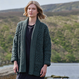 Woman wearing a green knitted Aran cardigan standing in a scenic outdoor setting with hills and water.