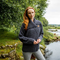 A woman with long red hair stands near a river, wearing a dark knitted Aran sweater with a large button. The background features lush greenery and a calm waterway, creating a serene outdoor setting.