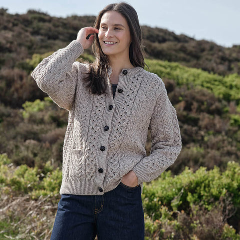 Person wearing a beige Women's Traditional Cable Knit Aran Cardigan, featuring intricate cable knit patterns and dark buttons, paired with dark blue jeans, standing outdoors with a natural landscape of greenery and hills in the background.