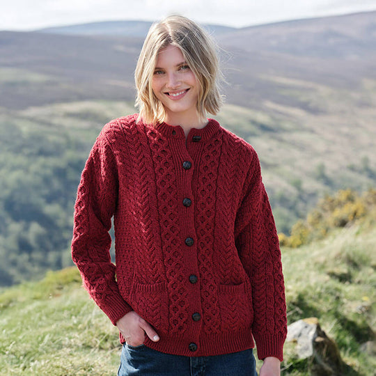 Woman wearing a red Womens Traditional Cable Knit Aran Cardigan standing in a scenic outdoor setting with mountains.