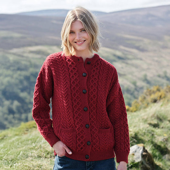 Woman wearing a red Womens Traditional Cable Knit Aran Cardigan standing in a scenic outdoor setting with mountains.