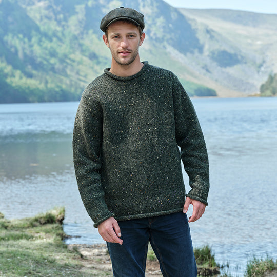 Man wearing a green sweater and flat cap standing by a lake with mountains in the background