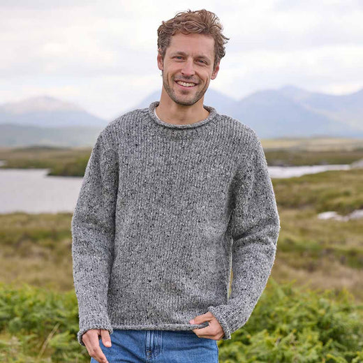 Man wearing a gray Men's Traditional Fisherman Irish Wool Sweater standing in a scenic outdoor setting with mountains and water.