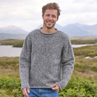 Man wearing a gray Men's Traditional Fisherman Irish Wool Sweater standing in a scenic outdoor setting with mountains and water.