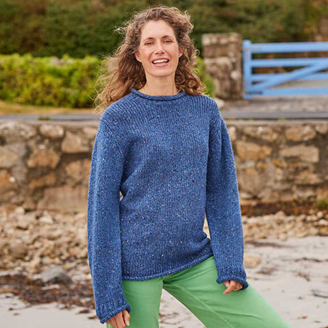 Woman wearing a blue Women's Traditional Fisherman Irish Wool Sweater standing outdoors with a stone wall and blue gate in the background.