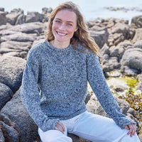 Woman wearing a grey Women's Traditional Fisherman Irish Wool Sweater sitting on rocky terrain by the sea.