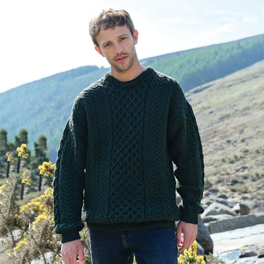 Man wearing a green Aran sweater standing in a scenic outdoor setting with mountains and rocks.