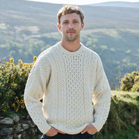 Man wearing a cream sweater standing in a scenic outdoor setting with mountains and greenery.