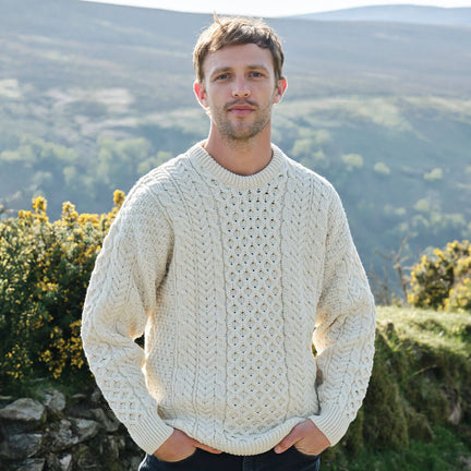 Man wearing a cream sweater standing in a scenic outdoor setting with mountains and greenery.