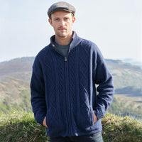 Man wearing a navy blue cable knit sweater and flat cap standing outdoors with mountains in the background