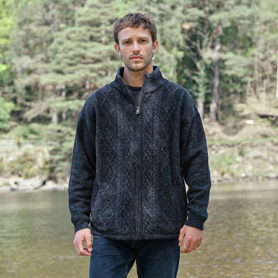 Man wearing a dark jacket standing by a body of water with trees in the background