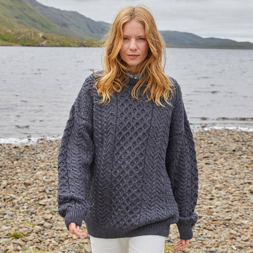Woman wearing a dark charcoal gray Aran sweater standing on a pebbly beach with water and mountains in the background.