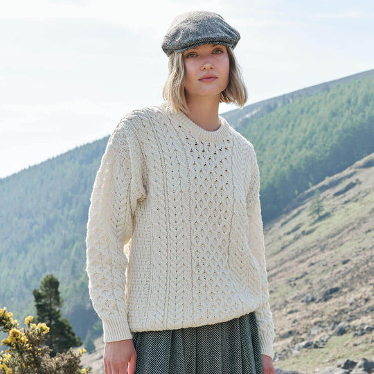 Woman wearing a cream knitted sweater and flat cap standing in a mountainous landscape